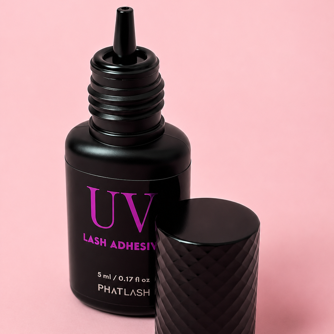 A bottle of PHATLASH UV Lash Adhesive in matte black with magenta lettering, shown next to its textured black cap on a solid pink background.